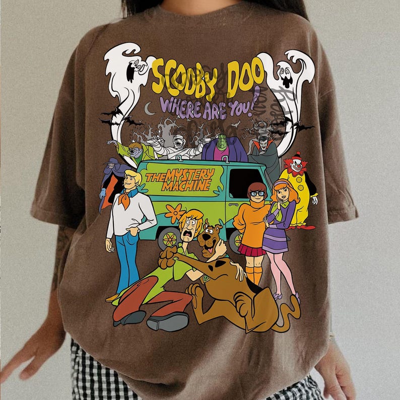 May include: A brown t-shirt featuring a graphic print of the Scooby-Doo cast, the Mystery Machine van, and ghosts. The text reads Scooby Doo Where Are You and The Mystery Machine. The design includes various characters like Shaggy, Velma, and Daphne.
