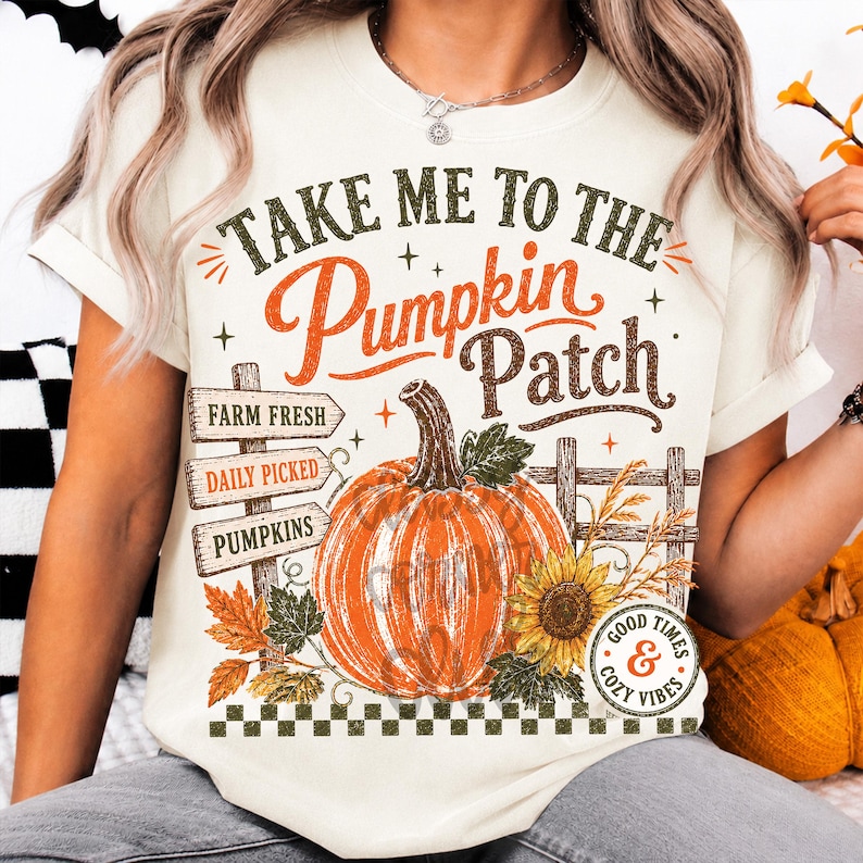 Pode incluir: Uma t-shirt cor de creme com uma estampa gr&aacute;fica com o texto "TAKE ME TO THE PUMPKIN PATCH". O design inclui uma ab&oacute;bora laranja grande, uma placa de madeira com "FARM FRESH", "DAILY PICKED", "PUMPKINS", um girassol e v&aacute;rias folhas.