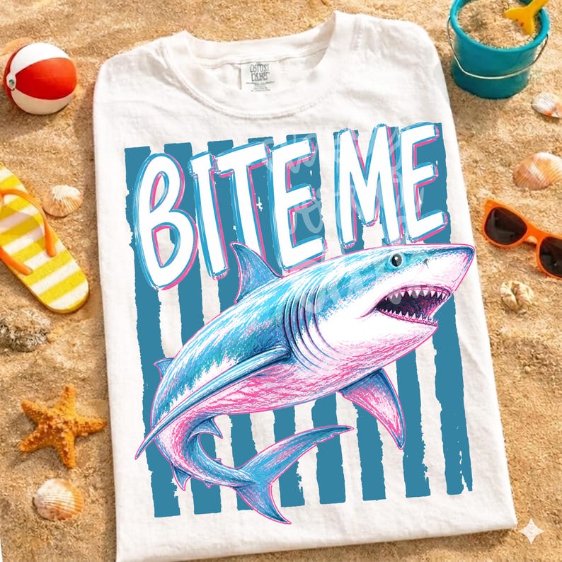 May include: White t-shirt featuring a blue and pink shark graphic with the text "BITE ME". The shark design has sharp teeth. The shirt is displayed on a sandy surface, surrounded by beach items.