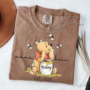 Może przedstawiać: Brązowa koszulka z nadrukiem kresk&oacute;wkowej postaci Winnie the Pooh trzymającego słoik miodu. Na koszulce widnieje napis "mama bear" i "EST. 2025".