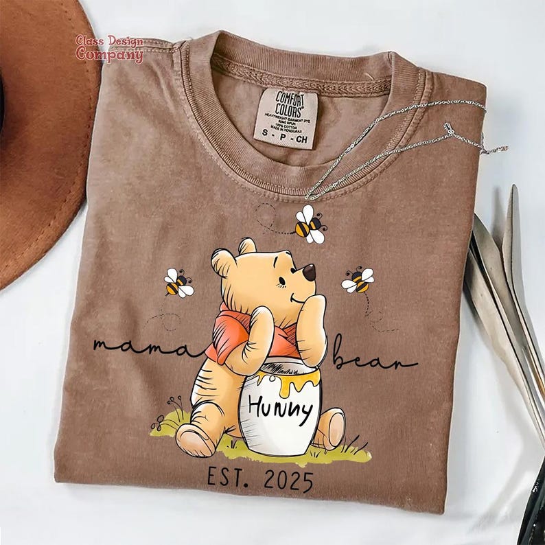 Może przedstawiać: Brązowa koszulka z nadrukiem kresk&oacute;wkowej postaci Winnie the Pooh trzymającego słoik miodu. Na koszulce widnieje napis "mama bear" i "EST. 2025".