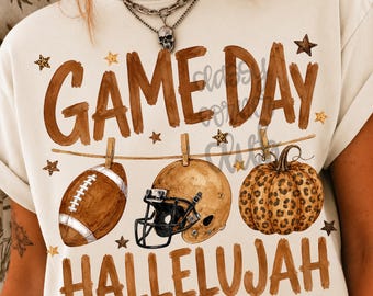 May include: A cream-colored t-shirt featuring the brown text GAME DAY HALLELUJAH. The graphic shows a football, a gold football helmet, and a leopard print pumpkin hanging from a clothesline, accented by small brown and leopard print stars.