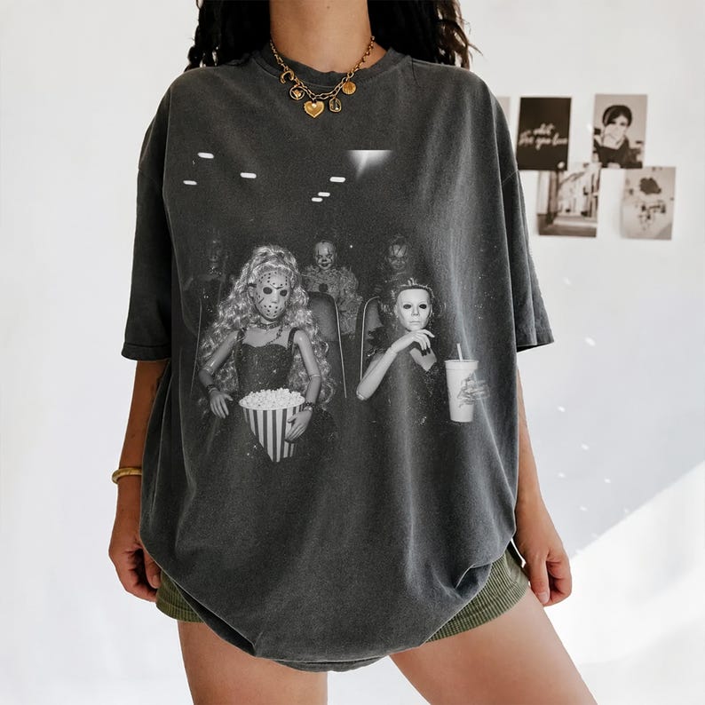 Puede incluir: Una camiseta gris carb&oacute;n de corte oversize con un estampado gr&aacute;fico en blanco y negro de mu&ntilde;ecas con m&aacute;scaras de pel&iacute;culas de terror sentadas en un cine con palomitas y una bebida. Tiene cuello redondo y mangas cortas.