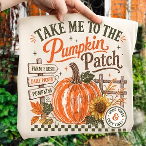 Pode incluir: Uma t-shirt de cor creme com uma impress&atilde;o gr&aacute;fica com o texto "TAKE ME TO THE Pumpkin Patch". O design inclui uma ab&oacute;bora laranja grande, uma placa de madeira com "FARM FRESH", "DAILY PICKED" e "PUMPKINS", um girassol e v&aacute;rias folhas.