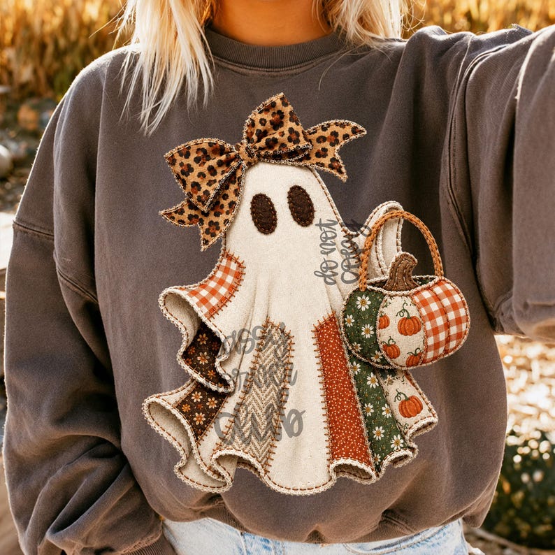 May include: A charcoal gray crewneck sweatshirt featuring a central graphic of a white ghost wearing a leopard print bow. The ghost is holding a pumpkin-shaped basket with orange gingham and floral patterns, and the ghost's body is composed of various fabric-like patches.
