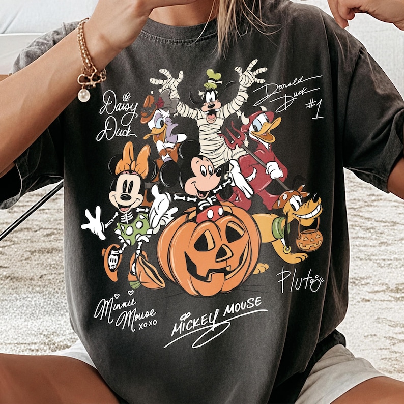 K&ouml;nnte beinhalten: Ein anthrazitfarbenes Kurzarm-T-Shirt mit einer Halloween-Grafik von Disney-Figuren, darunter Mickey Mouse in einem K&uuml;rbis, Minnie Mouse, Goofy als Mumie und Donald Duck als Teufel, erg&auml;nzt durch handschriftliche Namensz&uuml;ge.