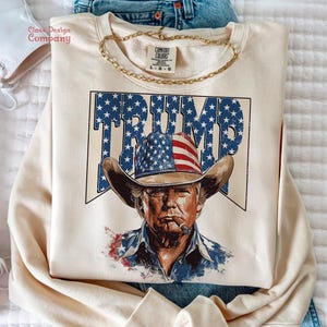 May include: A beige sweatshirt with a graphic print of a man wearing a cowboy hat with an American flag design. The text "TRUMP" is printed in blue and white stars on the hat.