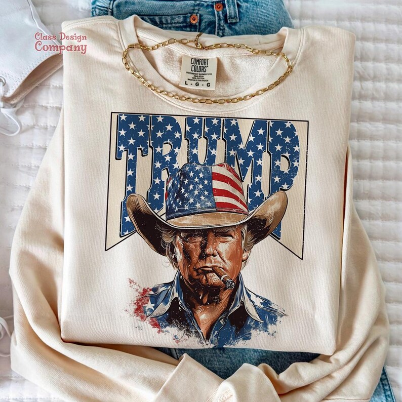 May include: A beige sweatshirt with a graphic print of a man wearing a cowboy hat with an American flag design. The text "TRUMP" is printed in blue and white stars on the hat.