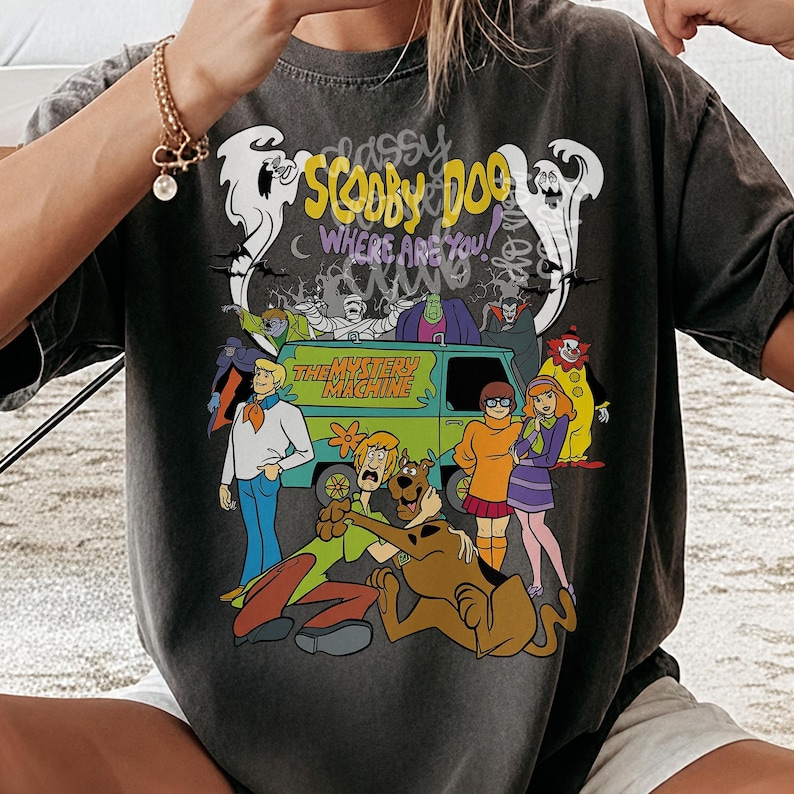 May include: A charcoal gray short-sleeve t-shirt featuring a graphic print of the Scooby-Doo cast, the Mystery Machine van, and various monsters. The text reads Scooby-Doo Where Are You and The Mystery Machine in colorful, bold lettering.