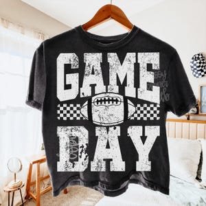 May include: A black short-sleeve t-shirt with a distressed white graphic print. The design features the text "GAME DAY" in large block letters with a football illustration and a checkered pattern strip positioned between the two words.