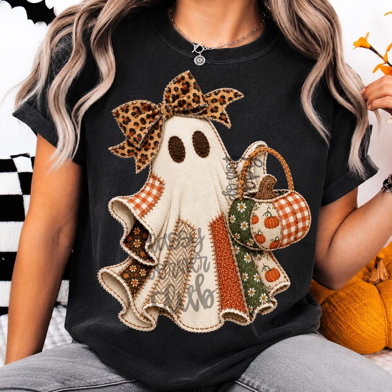 May include: A black t-shirt featuring a graphic of a ghost wearing a leopard print bow and holding a pumpkin basket. The ghost is decorated with various fabric patterns including orange plaid, brown floral, and herringbone textures with stitched edges.