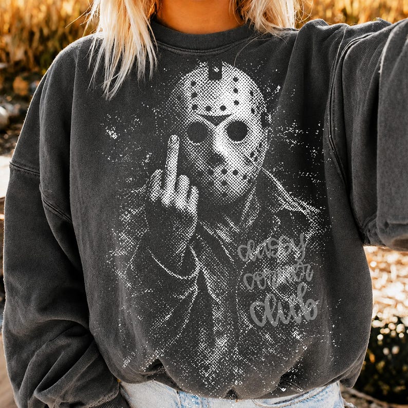 May include: A charcoal grey crewneck sweatshirt featuring a white distressed graphic of a hockey mask-wearing character giving the middle finger. The design includes the text "classy corner club" in a cursive script font on the lower right side.