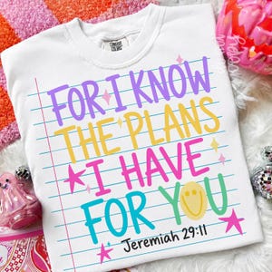 May include: A white t-shirt featuring a notebook paper background design with the text "FOR I KNOW THE PLANS I HAVE FOR YOU" in purple, yellow, pink, and teal, along with a yellow smiley face and small pink stars, plus "Jeremiah 29:11" below.