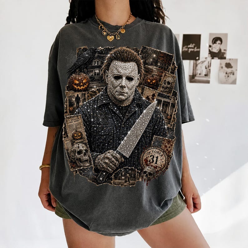 May include: A charcoal gray short-sleeve t-shirt featuring a large graphic print of a masked character holding a knife, surrounded by collage-style images of pumpkins, a skull, a raven, and a circular emblem with the text October 31.