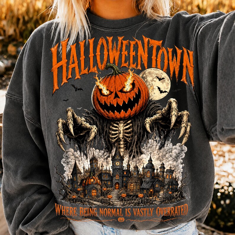 Puede incluir: Una sudadera gris carb&oacute;n con un gr&aacute;fico de una calabaza en llamas con brazos esquel&eacute;ticos extendi&eacute;ndose sobre un castillo oscuro. El texto dice HALLOWEENTOWN en la parte superior y WHERE BEING NORMAL IS VASTLY OVERRATED en la parte inferior.