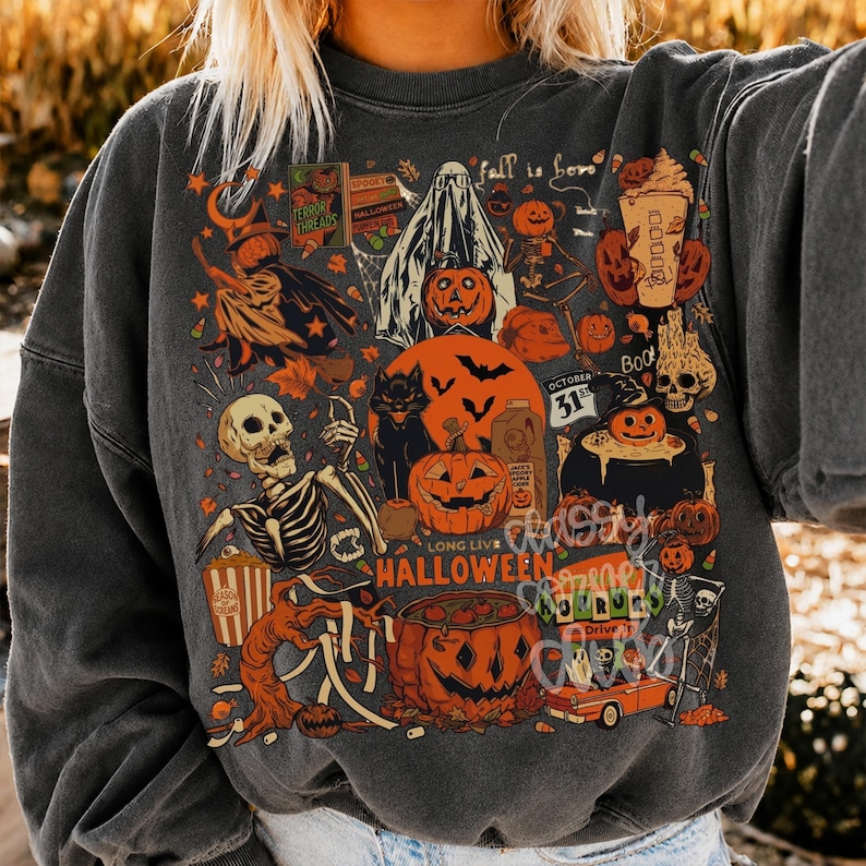 May include: A charcoal gray crewneck sweatshirt featuring a vibrant collage of Halloween-themed illustrations including a skeleton, a ghost, pumpkins, a black cat, a witch, a cauldron, and the text "Fall is here" and "Long Live Halloween".