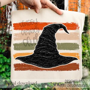 May include: A cream-colored tote bag featuring a black witch hat graphic centered over horizontal, distressed stripes in orange, beige, olive green, and brown. The text "classy corner club" is visible in a cursive font over the stripes.
