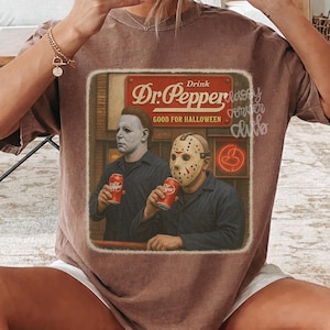 May include: A brown short-sleeve t-shirt featuring a graphic print of two horror movie characters holding red soda cans. The text reads Drink Dr. Pepper GOOD FOR HALLOWEEN. The design is framed in a rectangular border with a vintage aesthetic.