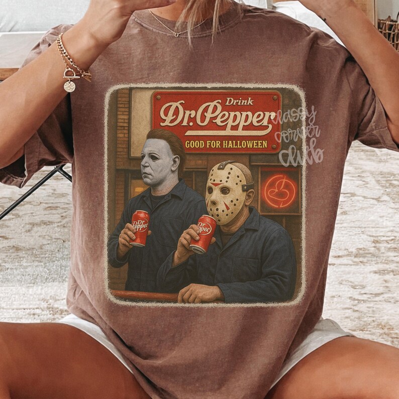 May include: A brown short-sleeve t-shirt featuring a graphic print of two horror movie characters holding red soda cans. The text reads Drink Dr. Pepper GOOD FOR HALLOWEEN. The design is framed in a rectangular border with a vintage aesthetic.