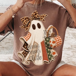 May include: A brown t-shirt featuring a graphic of a ghost wearing a leopard print bow. The ghost is composed of various fabric-like patches in orange, green, and brown patterns, and it is holding a small pumpkin-shaped basket with a handle.