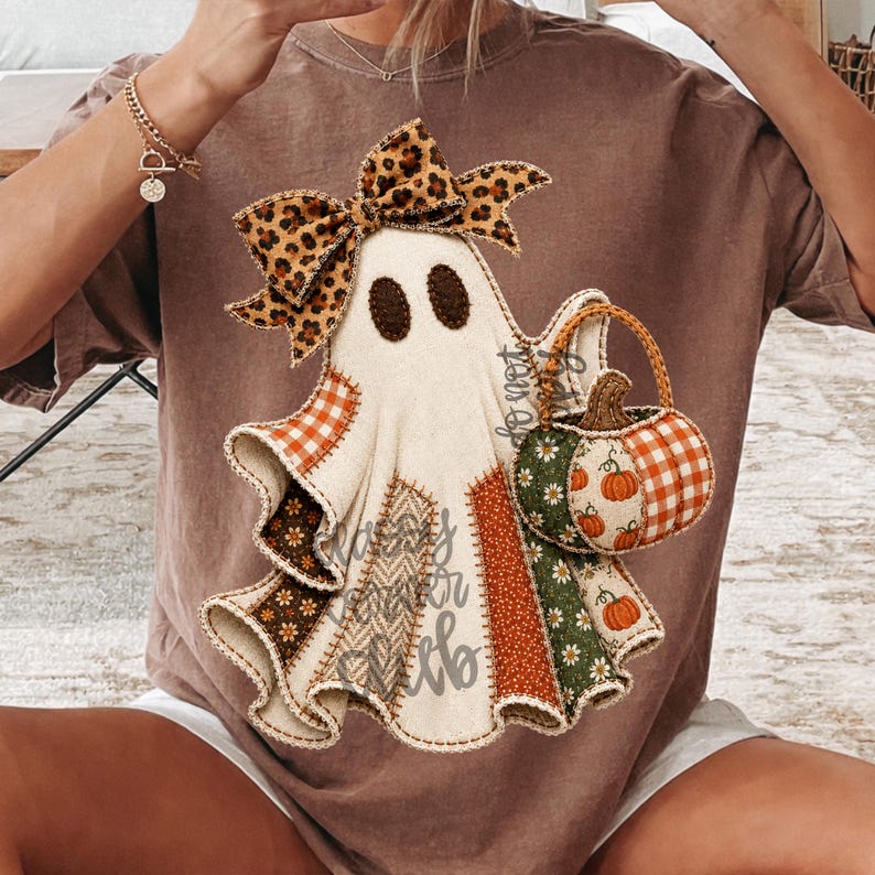 May include: A brown t-shirt featuring a graphic of a ghost wearing a leopard print bow. The ghost is composed of various fabric-like patches in orange, green, and brown patterns, and it is holding a small pumpkin-shaped basket with a handle.