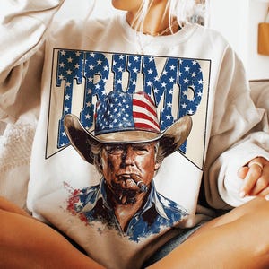May include: A white sweatshirt with a graphic print of a man wearing a cowboy hat with an American flag design. The text "TRUMP" is printed in large letters above the man's head.