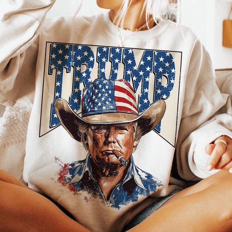 May include: A white sweatshirt with a graphic print of a man wearing a cowboy hat with an American flag design. The text "TRUMP" is printed in large letters above the man's head.