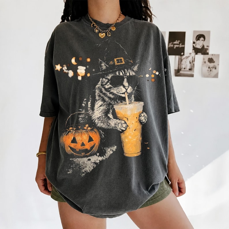 Puede incluir: Una camiseta gris carb&oacute;n de manga corta con un estampado gr&aacute;fico de un gato con sombrero de bruja y gafas de sol, sosteniendo un caf&eacute; helado. Junto al gato hay un cubo de calabaza naranja, con peque&ntilde;as estrellas y lunas arriba.