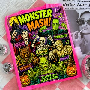 May include: A bright pink hoodie featuring a colorful graphic print titled Monster Mash. The design displays illustrations of Dracula, Frankenstein, a skeleton, a mummy, a werewolf, and a creature from the black lagoon, along with two jack-o'-lanterns.
