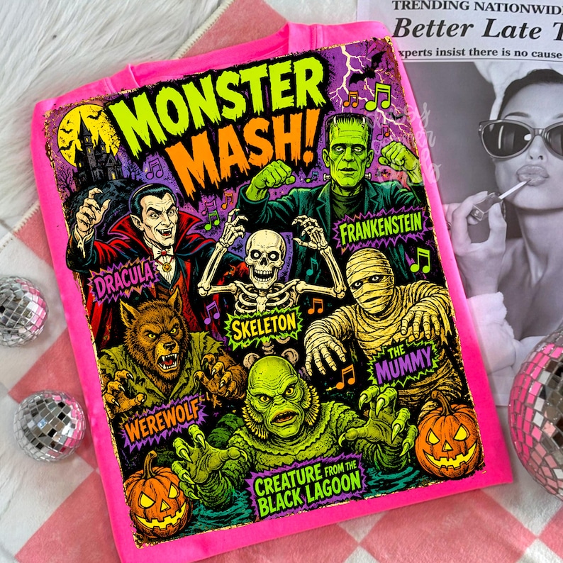 May include: A bright pink hoodie featuring a colorful graphic print titled Monster Mash. The design displays illustrations of Dracula, Frankenstein, a skeleton, a mummy, a werewolf, and a creature from the black lagoon, along with two jack-o'-lanterns.