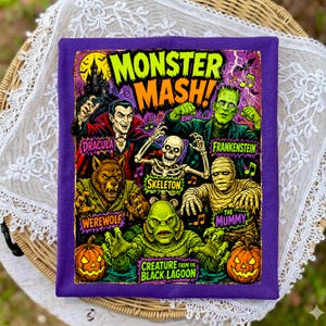 May include: A purple t-shirt featuring a graphic print titled Monster Mash with illustrations of Dracula, Frankenstein, a skeleton, a werewolf, the mummy, and the creature from the black lagoon, surrounded by musical notes and two jack-o'-lanterns.