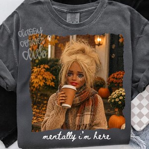 May include: A charcoal gray t-shirt featuring a graphic print of a person with blonde hair wearing a beige sweater and scarf, holding a coffee cup. The background shows autumn foliage and pumpkins. The text reads, "mentally i'm here."