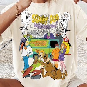 May include: A cream-colored t-shirt featuring a graphic print of the Scooby-Doo cast, the Mystery Machine van, and various monsters. The text reads Scooby Doo Where Are You and The Mystery Machine. The design includes ghosts, bats, and trees.
