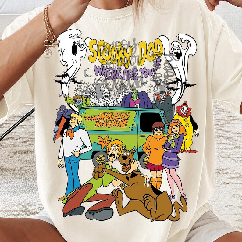 May include: A cream-colored t-shirt featuring a graphic print of the Scooby-Doo cast, the Mystery Machine van, and various monsters. The text reads Scooby Doo Where Are You and The Mystery Machine. The design includes ghosts, bats, and trees.