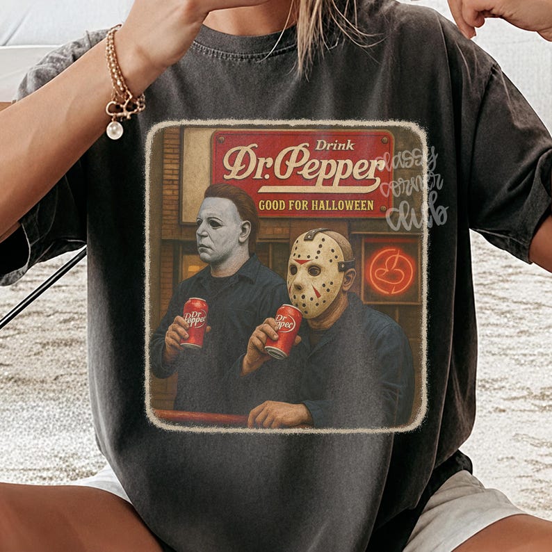 May include: A charcoal gray t-shirt featuring a graphic print of two horror movie characters holding red Dr. Pepper cans. The text reads Drink Dr. Pepper Good for Halloween. The design has a distressed, vintage-inspired square border.