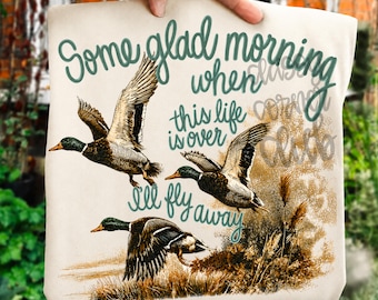 May include: A cream-colored t-shirt featuring a graphic print of three mallard ducks in flight and resting near reeds. The text reads, "Some glad morning when this life is over I'll fly away," written in a dark teal cursive script font.