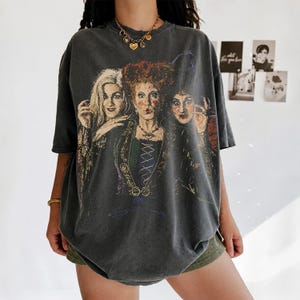 Puede incluir: Una camiseta de manga corta en color gris carb&oacute;n con un gran estampado gr&aacute;fico a color de tres mujeres con peinados distintivos y ropa de &eacute;poca. La prenda presenta un corte holgado y extragrande con cuello redondo y un acabado desgastado.
