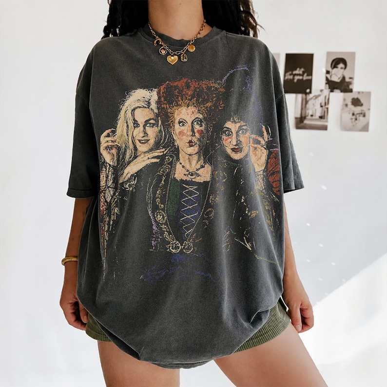 Puede incluir: Una camiseta de manga corta en color gris carb&oacute;n con un gran estampado gr&aacute;fico a color de tres mujeres con peinados distintivos y ropa de &eacute;poca. La prenda presenta un corte holgado y extragrande con cuello redondo y un acabado desgastado.