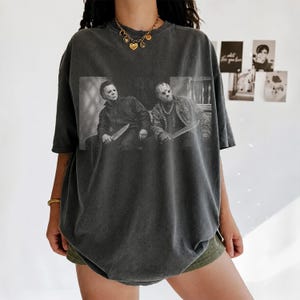 Puede incluir: Una camiseta gris carb&oacute;n de corte oversize con un estampado gr&aacute;fico en blanco y negro de dos personajes de pel&iacute;culas de terror, Michael Myers y Jason Voorhees, cada uno sosteniendo un cuchillo. La prenda tiene cuello redondo y mangas cortas.