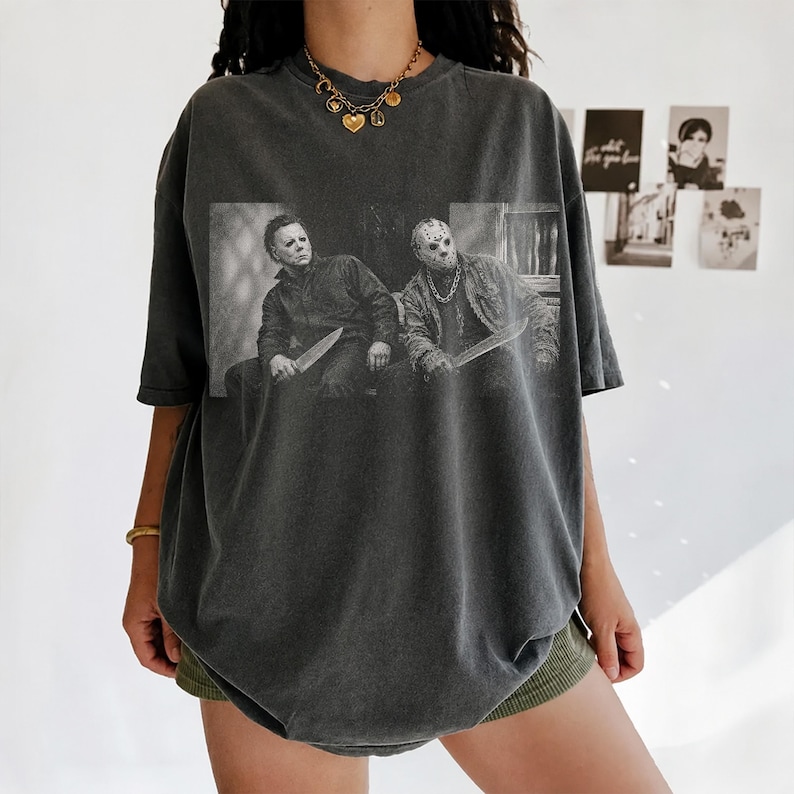 Puede incluir: Una camiseta gris carb&oacute;n de corte oversize con un estampado gr&aacute;fico en blanco y negro de dos personajes de pel&iacute;culas de terror, Michael Myers y Jason Voorhees, cada uno sosteniendo un cuchillo. La prenda tiene cuello redondo y mangas cortas.