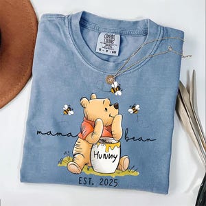 Może przedstawiać: Jasnoniebieska koszulka z nadrukiem kresk&oacute;wkowego obrazka Winnie the Pooh trzymającego słoik miodu. Na koszulce widnieje napis "mama bear" i "EST. 2025".
