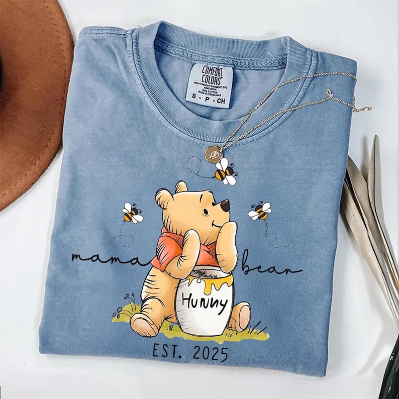 Może przedstawiać: Jasnoniebieska koszulka z nadrukiem kresk&oacute;wkowego obrazka Winnie the Pooh trzymającego słoik miodu. Na koszulce widnieje napis "mama bear" i "EST. 2025".
