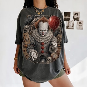 May include: A charcoal gray t-shirt featuring a graphic print of a clown with a red balloon, skulls, a raven, and tickets labeled ADMIT ONE and OCTOBER 31. The design includes intricate details of the clown's ruffled collar and red buttons.