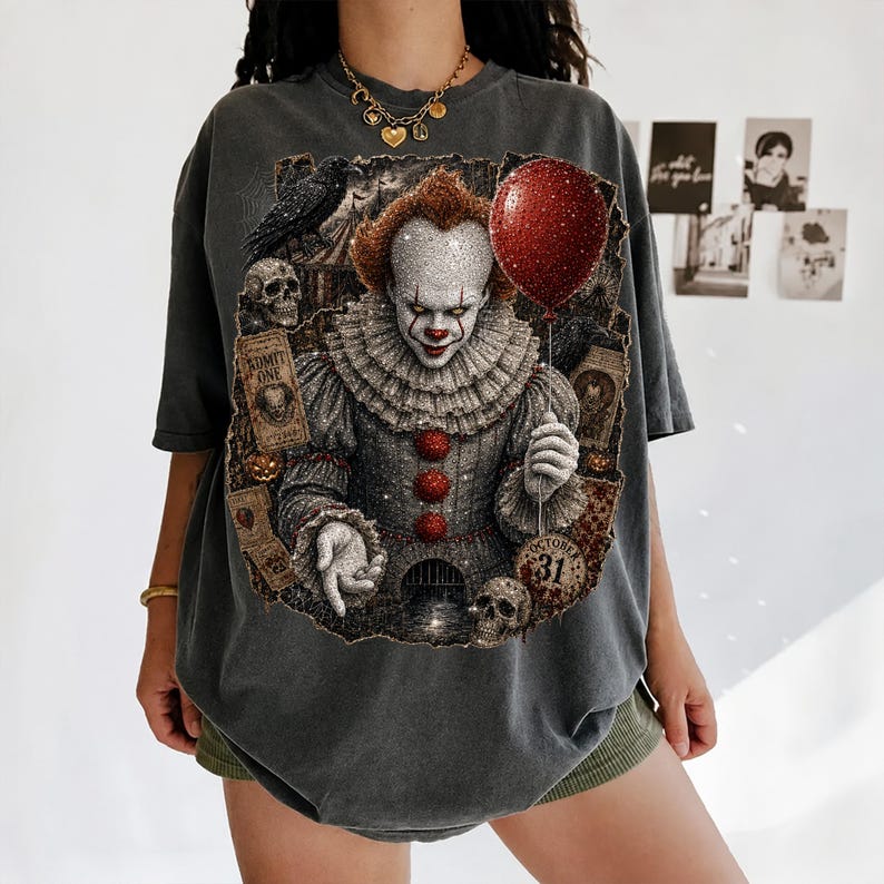 May include: A charcoal gray t-shirt featuring a graphic print of a clown with a red balloon, skulls, a raven, and tickets labeled ADMIT ONE and OCTOBER 31. The design includes intricate details of the clown's ruffled collar and red buttons.