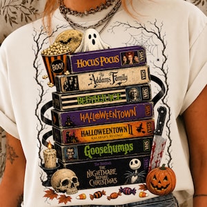May include: A white t-shirt featuring a graphic print of a stack of VHS tapes titled Hocus Pocus, The Addams Family, Beetlejuice, Halloweentown, Halloweentown II, Goosebumps, and The Nightmare Before Christmas, surrounded by a ghost, skull, and pumpkin.