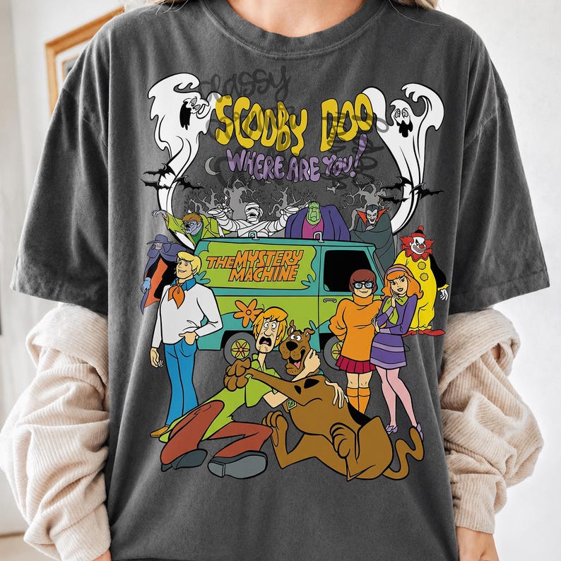 May include: A charcoal gray t-shirt featuring a graphic print of the Scooby-Doo Where Are You! cast, including Fred, Shaggy, Scooby-Doo, Velma, and Daphne standing in front of The Mystery Machine van, surrounded by various ghosts and monsters.