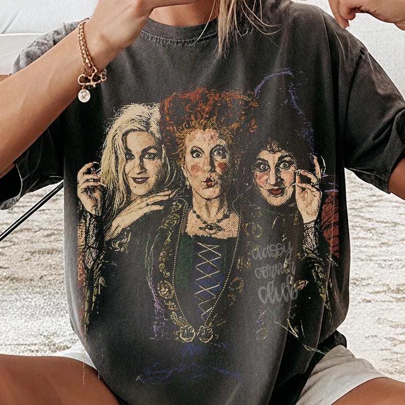 Peut inclure: Un t-shirt gris anthracite &agrave; manches courtes avec une impression graphique repr&eacute;sentant trois femmes aux coiffures distinctes et aux v&ecirc;tements sombres. La figure centrale a des cheveux roux vifs, tandis que les autres ont des cheveux blonds et fonc&eacute;s.