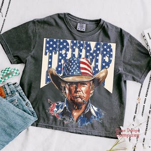 May include: A gray t-shirt with a vintage-style graphic of a man wearing a cowboy hat and a red, white, and blue bandana. The graphic says "TRUMP" in large letters.