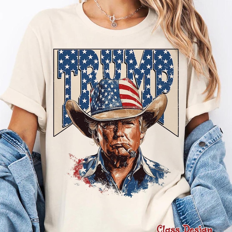 May include: A white t-shirt with a graphic of a man wearing a cowboy hat with an American flag design. The text "TRUMP" is written in large letters above the man's head.
