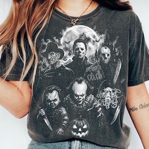 May include: A charcoal gray t-shirt featuring a white graphic print of five iconic horror movie characters, including a masked slasher, a clown, and a doll, set against a full moon background with bats and a jack-o'-lantern at the bottom.